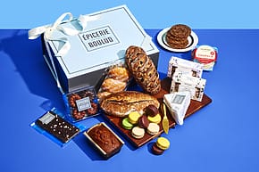 Gift box with assorted breads, macarons, cookies, cheese, and chocolate from Épicerie Boulud on a blue background.