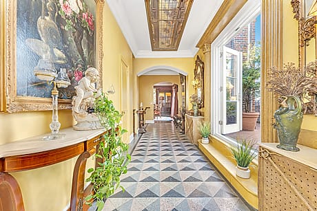 Elegant hallway with tiled floor, yellow walls, framed paintings, green plants, and ornate sculptures next to large windows.