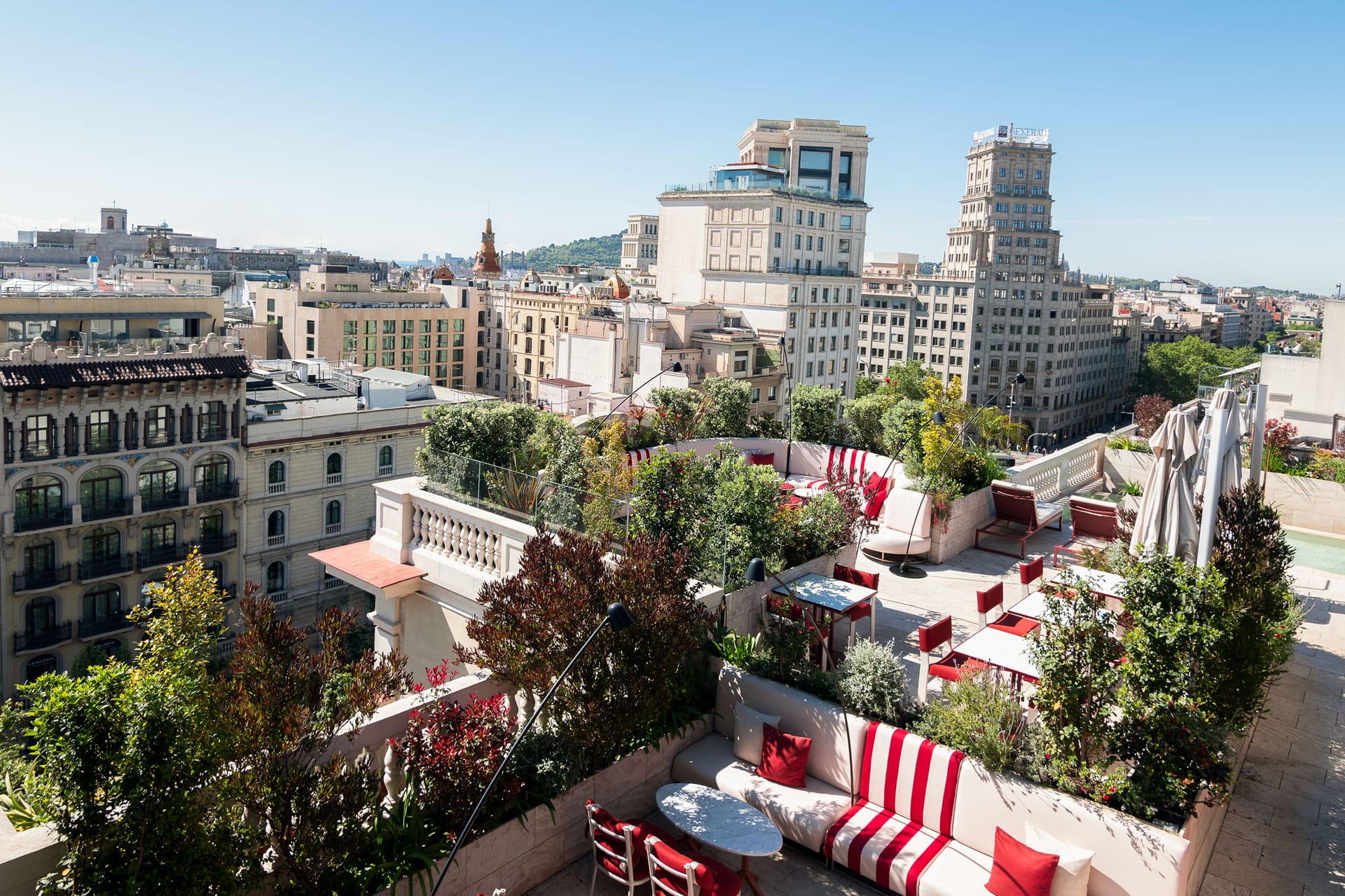 Rooftop terrace with red and white furniture, overlooking a cityscape with historic buildings and greenery in the background.