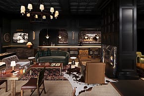 Chic bar interior with vintage furniture, patterned rugs, leather chairs, chandeliers, and framed photos on dark walls.