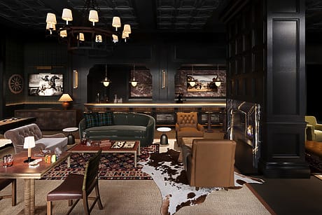 Chic bar interior with vintage furniture, patterned rugs, leather chairs, chandeliers, and framed photos on dark walls.