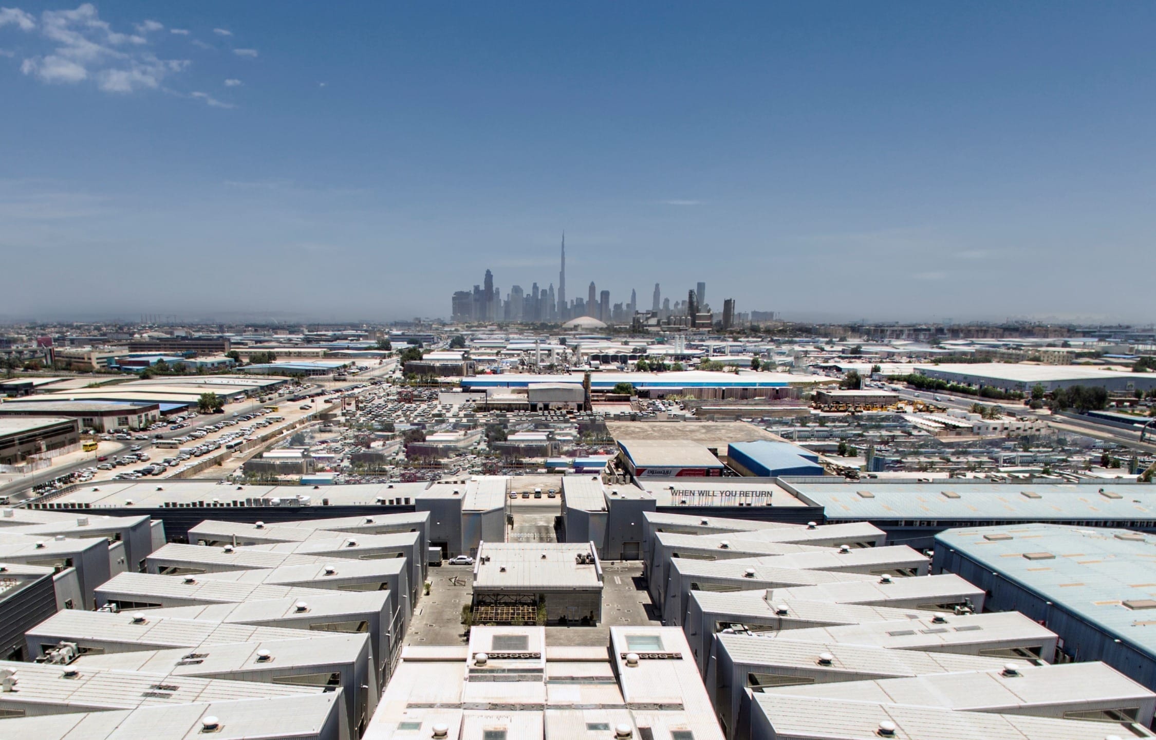 Aerial view of an industrial area with city skyline in the distance under a clear blue sky.