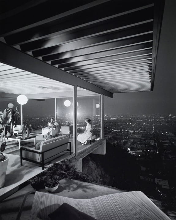 Modern glass-walled house at night overlooking city lights, with people relaxing on sofas inside.