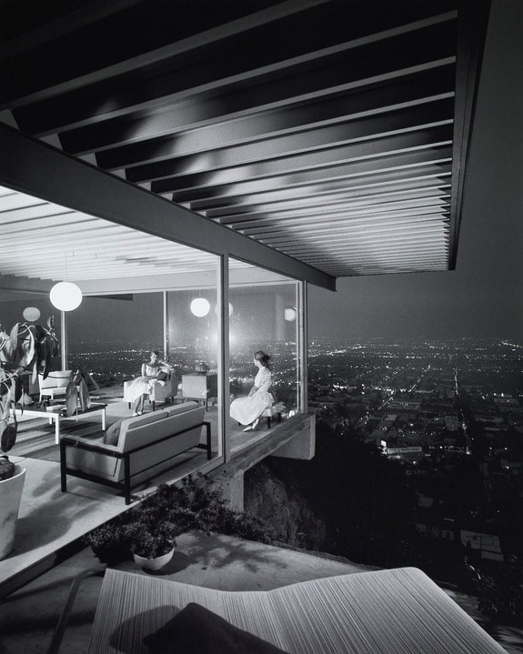 Modern glass-walled house at night overlooking city lights, with people relaxing on sofas inside.