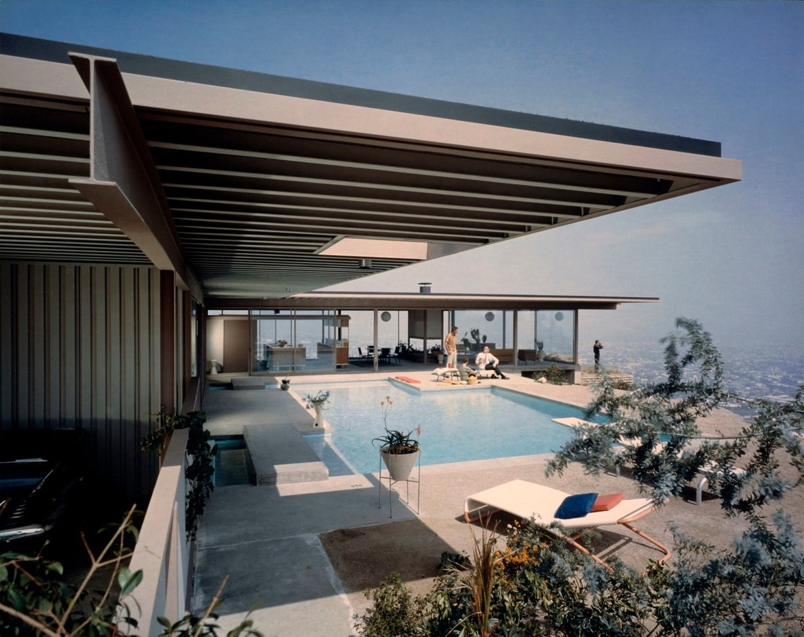 Modernist house with open-air pool, people relaxing by the water, overlooking city skyline under a clear blue sky.