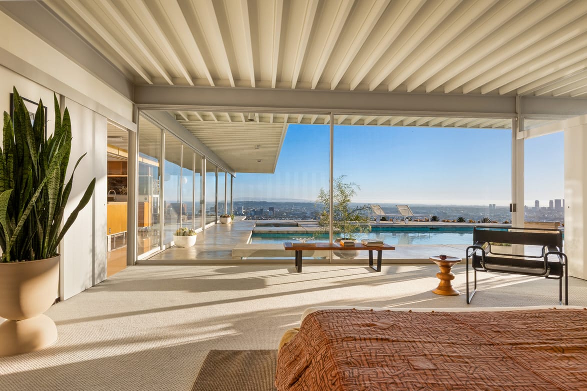Modern bedroom with large windows overlooking a pool and city skyline, with a potted plant and stylish decor elements.