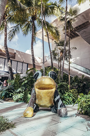 Large whimsical frog sculpture surrounded by tropical plants in outdoor urban setting with palm trees and modern architecture
