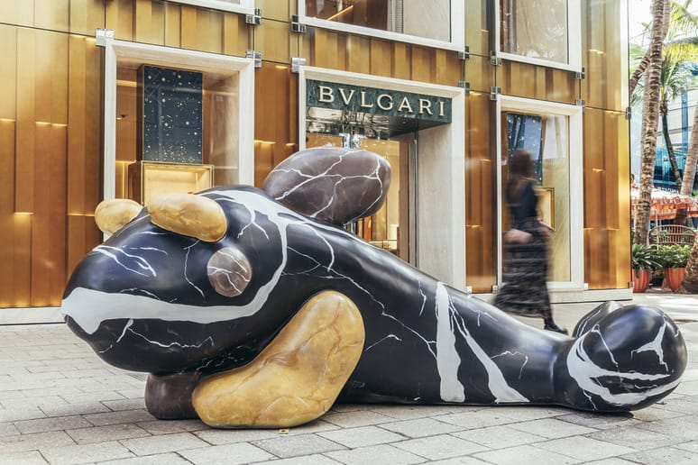 Large abstract sculpture resembling an animal in front of Bulgari store, blurred pedestrian walking past on a sunny day