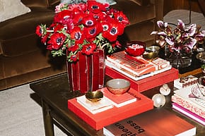 Coffee table with red and black books, colorful bowls, and a vase of vibrant red flowers in a cozy living room setting.