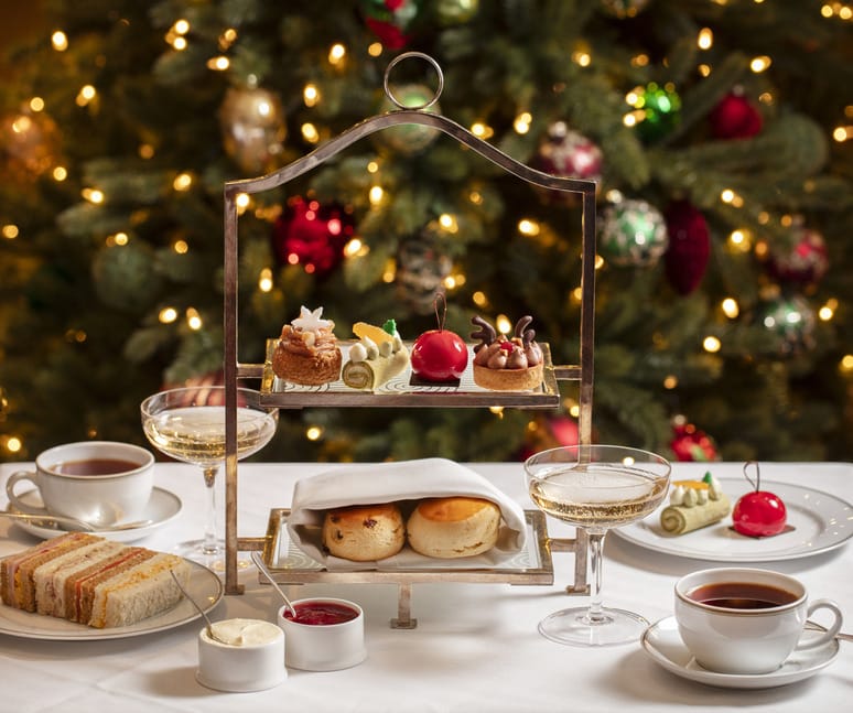 Festive afternoon tea setup with pastries, sandwiches, scones, and tea cups in front of a decorated Christmas tree.