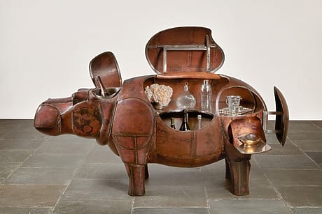 Copper sculpture of a hippopotamus functioning as a bar cabinet, displaying glasses and decanters inside on a tile floor.