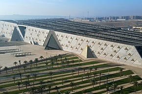 Aerial view of the Grand Egyptian Museum with geometric design, palm trees, and cityscape in the background.