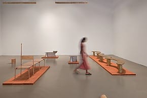Blurred person walking through minimalist art gallery with modern sculptures and furniture on display.