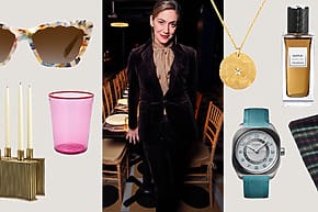 Woman in a velvet suit surrounded by luxury accessories, including sunglasses, candles, drinkware, perfume, jewelry, and a watch.