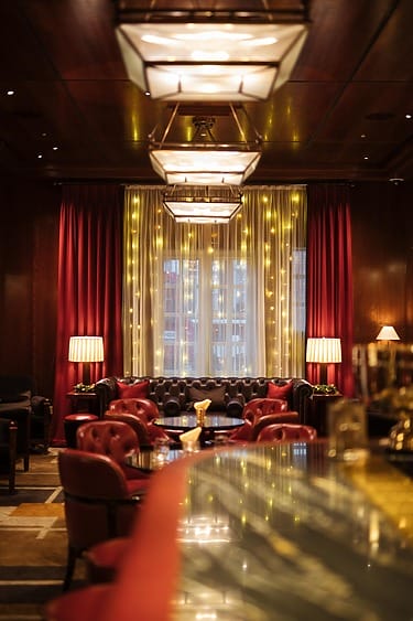 Cozy lounge with leather chairs, soft lighting, red curtains, and a glowing window creating an inviting atmosphere.