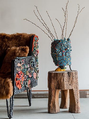 Colorful textured chair and vase on a rustic wooden stool with branches, set against a neutral wall background.