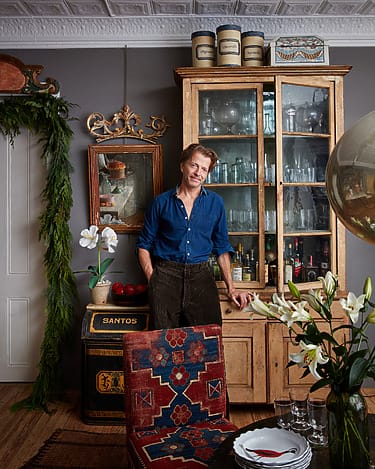 Man in a blue shirt leaning on a wooden cabinet with glassware, surrounded by decorative items and blooming lilies in a cozy room.