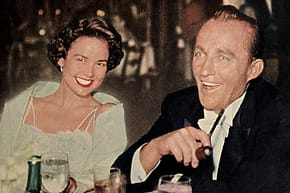 A man and woman smiling while seated at a dinner table, surrounded by glasses and plates in a warmly lit setting.