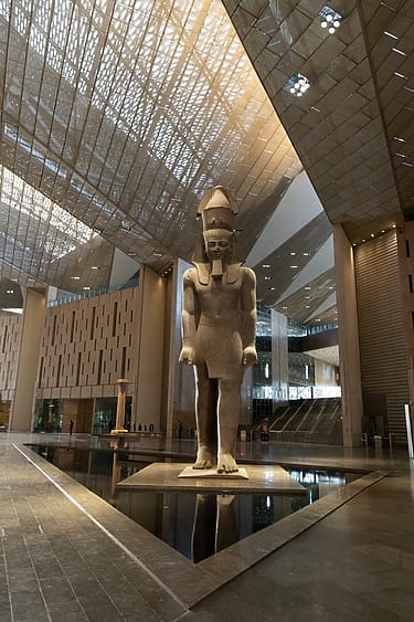 Colossal ancient statue in a modern museum hall with a reflective water feature and intricate ceiling design.