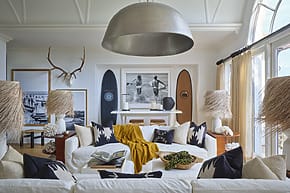 Coastal themed living room with surfboards, large pendant lamp, white sofa, decorative pillows, and antler wall decor.