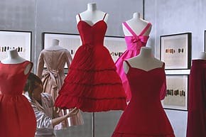 Designer adjusting red and pink dresses on mannequins in a fashion studio with framed sketches on the walls.