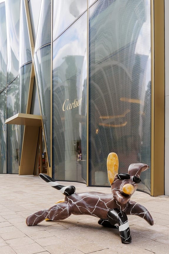 Sculpture of a reclining figure outside a modern Cartier store with tall glass windows and golden accents.