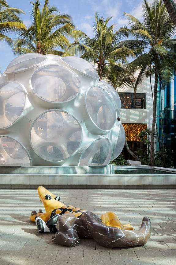 Sculpture of a yellow alligator on pavement with a futuristic geodesic dome and palm trees in the background.
