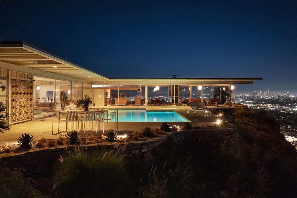 Modern hillside house with large windows, a lit swimming pool, and a view of a city skyline at night.