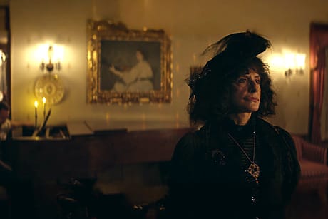 Woman in Victorian attire stands in dimly lit room with a painting and candles in the background.