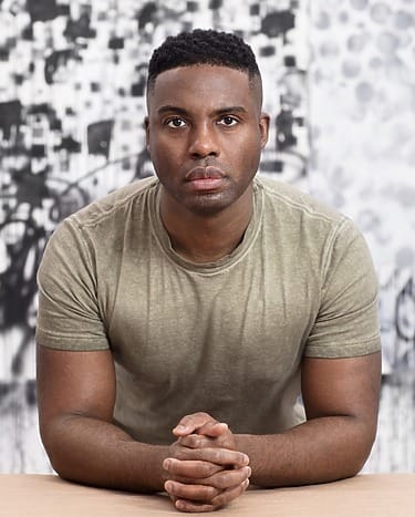 Person sitting with hands clasped, wearing a green shirt, in front of a black and white abstract background.
