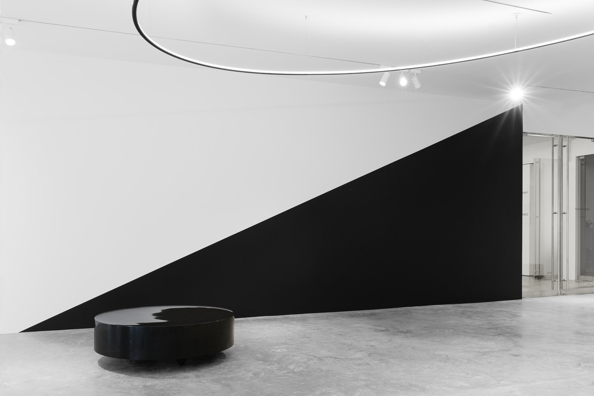 Modern art gallery interior with a black and white geometric wall design and a round black bench on polished concrete floor.