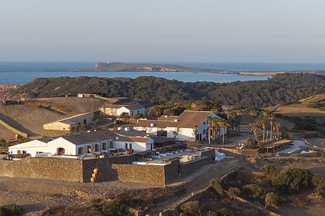 Rural estate overlooking the sea with white buildings, palm trees, and distant islands under a clear sky at sunset.