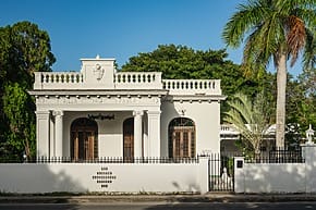 Historic white villa with arched windows and ornate detailing, surrounded by lush greenery and a palm tree.