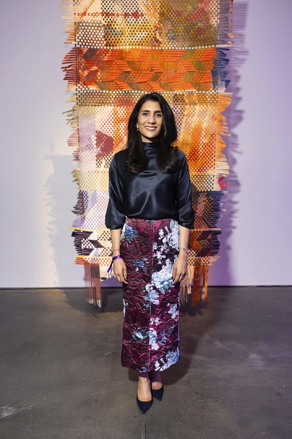 Person standing in front of a colorful abstract art piece, wearing a satin blouse and floral skirt, indoor setting.