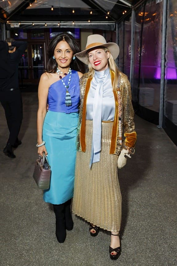 Two women posing at an event, one wearing a blue dress and the other in a gold skirt with a hat, both smiling.