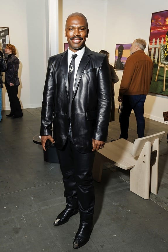 Man in black leather suit and boots standing at an art gallery event.