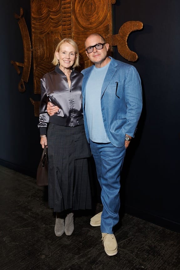 A smiling couple standing together, the woman in a satin shirt and skirt, the man in a blue suit with a casual stance.