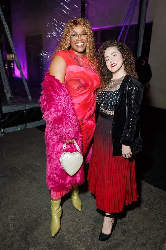 Two people smiling at a nighttime event, with vibrant outfits and a heart-shaped purse, standing indoors with pink lighting.