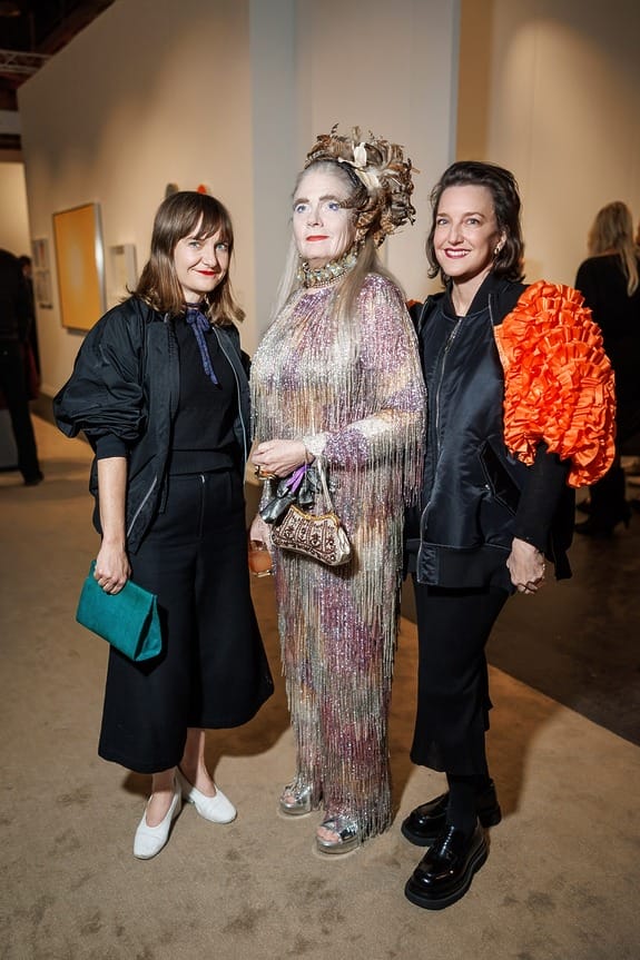 Three women posing at an art gallery event, one in a glittering costume, another with a vibrant shoulder accessory