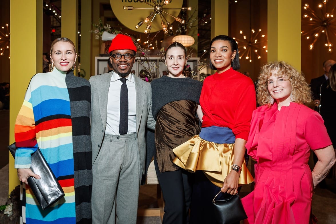 Group of five people smiling, wearing colorful and stylish outfits at a social event, with decorative lighting in the background.