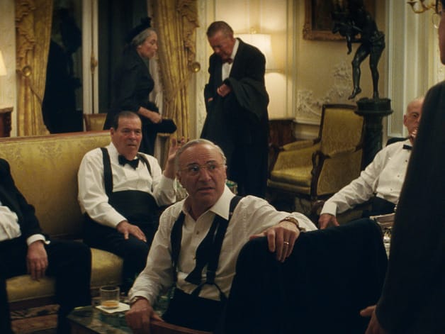 Group of people in formal attire engage in a serious discussion in an elegantly furnished room with ornate decor.