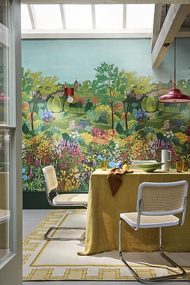 Colorful dining room with mural of vibrant garden, yellow tablecloth, wicker chairs, red light fixture, large windows.