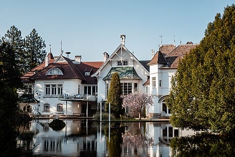 Large white mansion with red roof reflecting on tranquil pond surrounded by trees and clear blue sky.