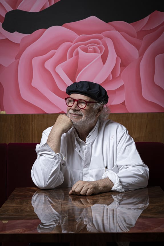 Man in a white shirt and black beret sitting at a table with a pink rose mural in the background