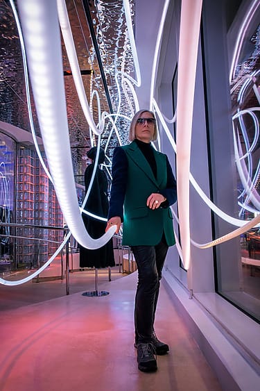 Person in stylish outfit stands in neon-lit room with reflective surfaces, creating a futuristic and artistic ambiance.