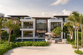 Modern beachfront villa with pool, surrounded by palm trees under a blue sky.