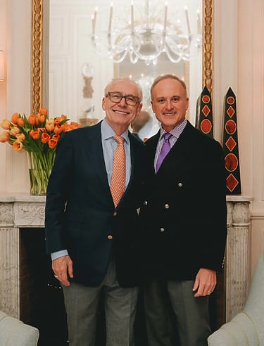 Two smiling men standing together in a well-decorated room with a marble fireplace and a vase of orange tulips.