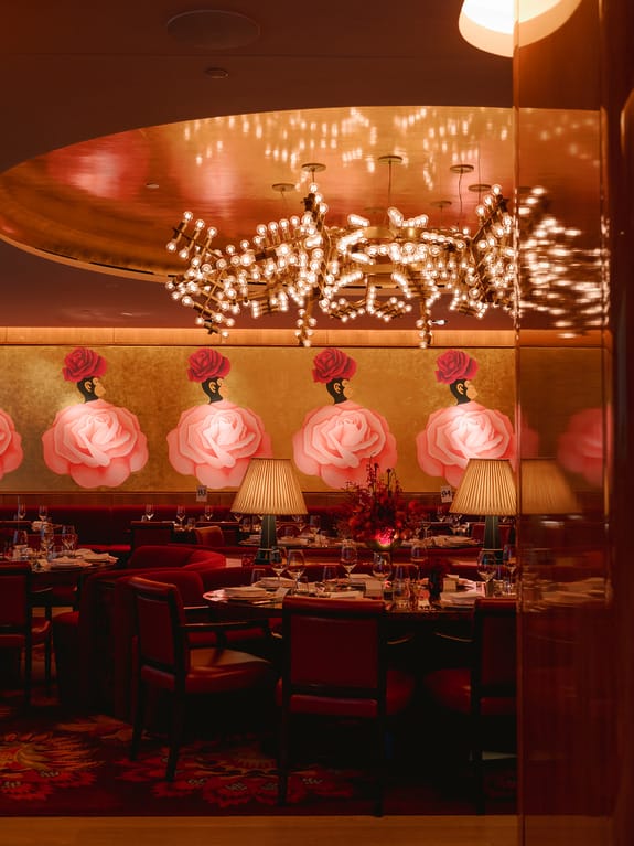 Elegant restaurant interior with rose artwork, plush seating, and a sparkling chandelier above neatly set dining tables.