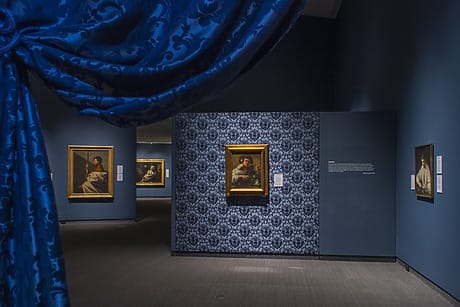 Interior view of an art gallery with framed paintings on blue patterned walls and a blue curtain in the foreground.