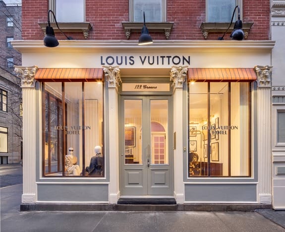 Storefront of a Louis Vuitton boutique with large windows and mannequins on display inside located on a city street corner.